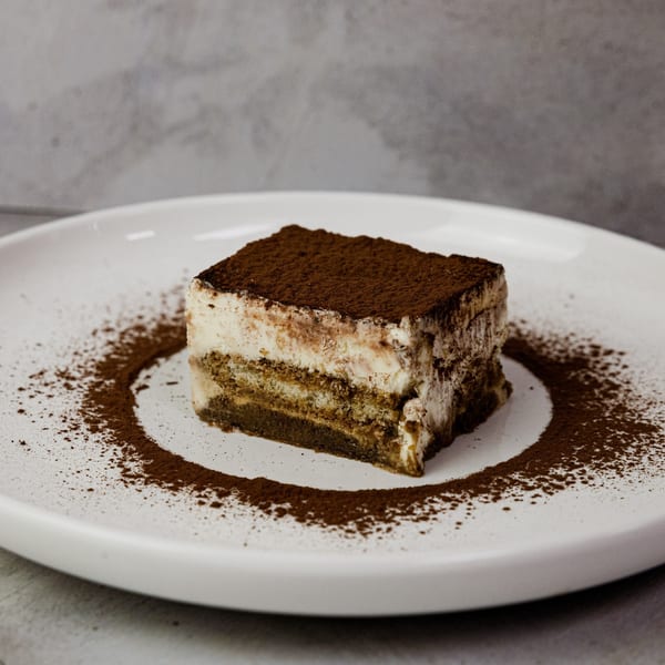 A slice of tiramisu dessert, dusted with cocoa powder, on a white plate.