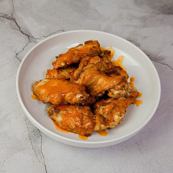 A white bowl filled with golden-brown chicken wings generously coated in a vibrant orange buffalo sauce.