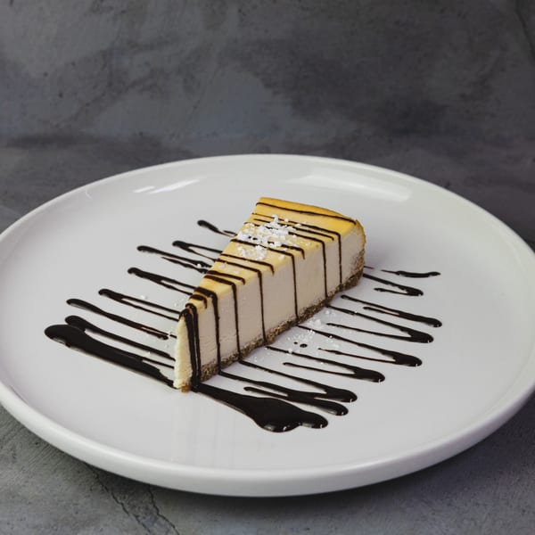 A slice of cheesecake with chocolate drizzle and powdered sugar on a white plate.